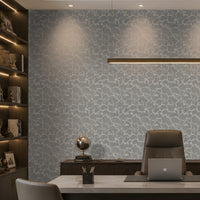 A modern home office featuring a light gray and white abstract patterned wallpaper on the main wall, a dark wood built-in bookshelf with warm lighting on the left, a white desk, a brown leather office chair, and a laptop on a desk mat.