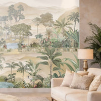A beige-toned living room with a large wall mural featuring palm trees, tropical plants, and a serene landscape with water and distant hills. In front of the mural is a light beige sofa with textured cushions, a side table with a gold-based lamp, and a tall indoor palm plant adding greenery to the neutral setting.