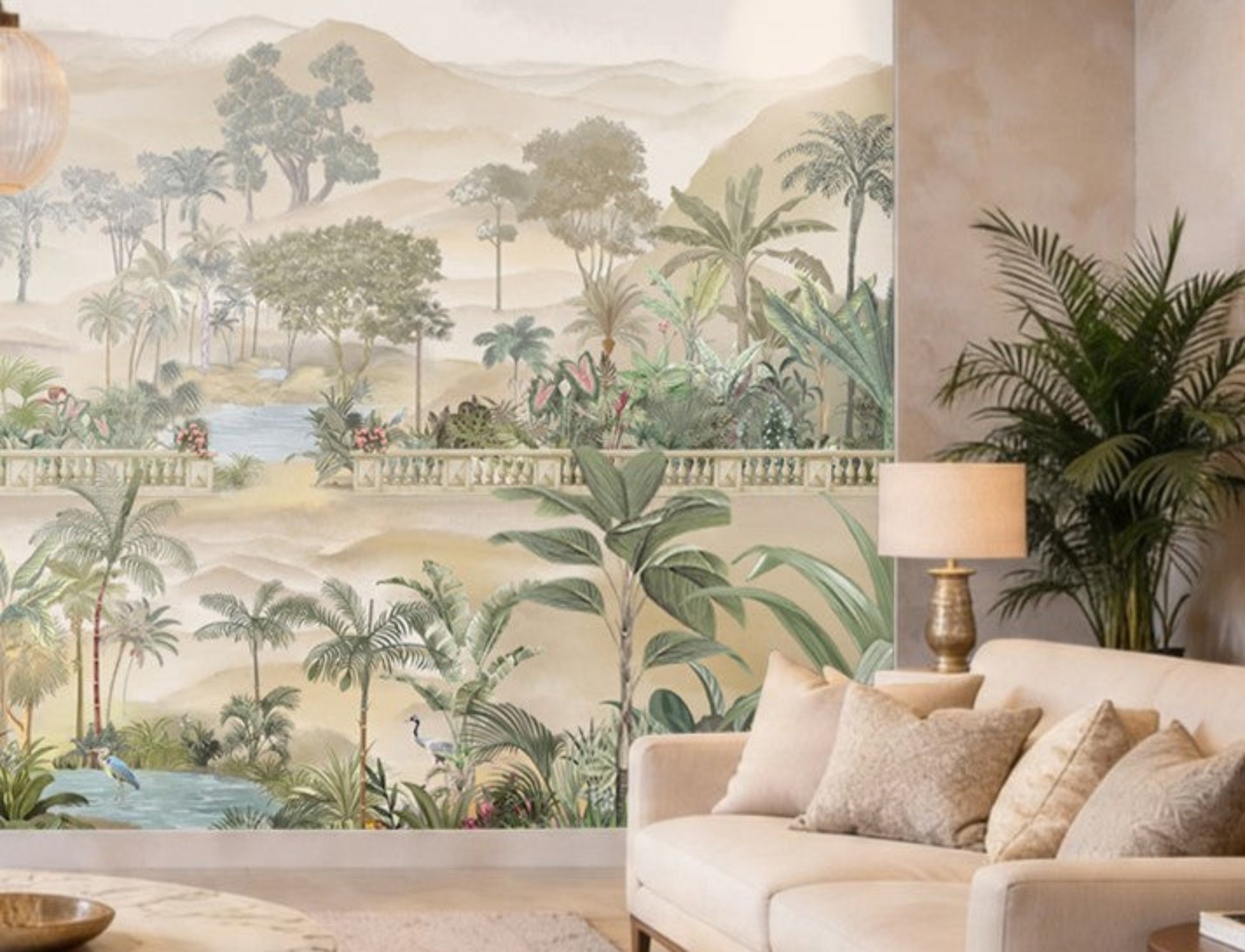 A beige-toned living room with a large wall mural featuring palm trees, tropical plants, and a serene landscape with water and distant hills. In front of the mural is a light beige sofa with textured cushions, a side table with a gold-based lamp, and a tall indoor palm plant adding greenery to the neutral setting.