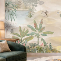 A beige-toned living room with a large wall mural featuring palm trees, tropical plants, and a serene landscape with water and distant hills. In front of the mural is a light beige sofa with textured cushions, a side table with a gold-based lamp, and a tall indoor palm plant adding greenery to the neutral setting.