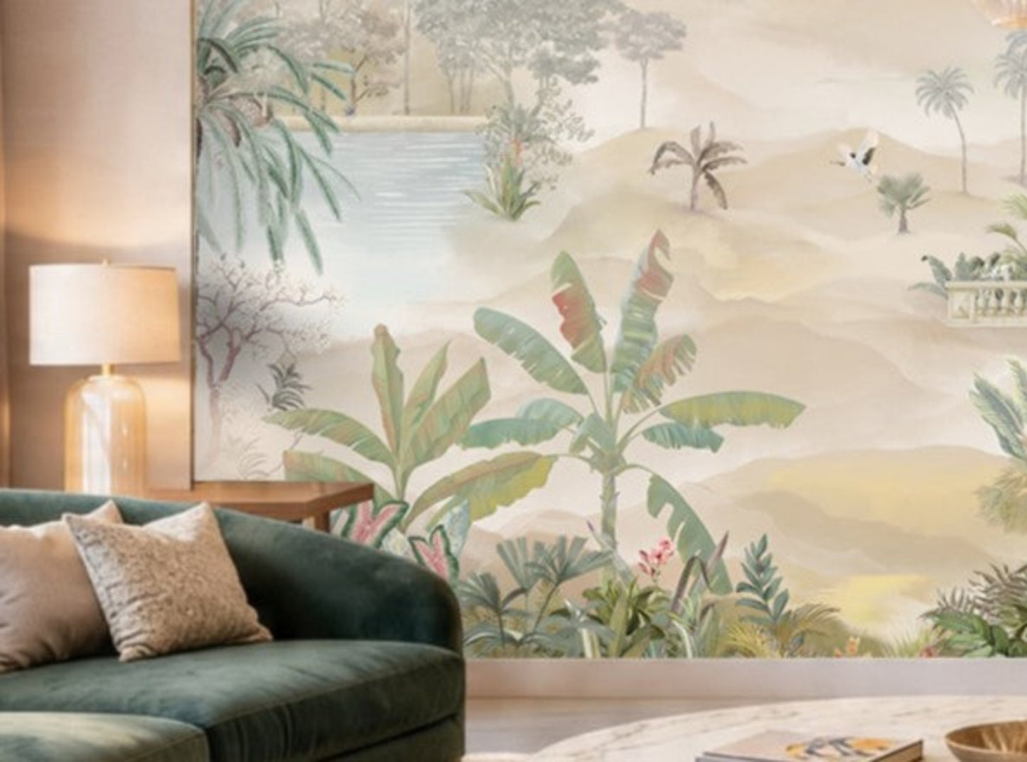 A beige-toned living room with a large wall mural featuring palm trees, tropical plants, and a serene landscape with water and distant hills. In front of the mural is a light beige sofa with textured cushions, a side table with a gold-based lamp, and a tall indoor palm plant adding greenery to the neutral setting.