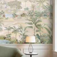 A spacious living room in soft beige tones featuring a large wall mural of palm trees, tropical plants, and a serene landscape with water and distant hills. The room includes a beige sofa with cushions, a green velvet sofa, two wooden armchairs, and a round marble coffee table. Warm pendant lights hang from the ceiling, and indoor plants complement the calm, neutral setting.
