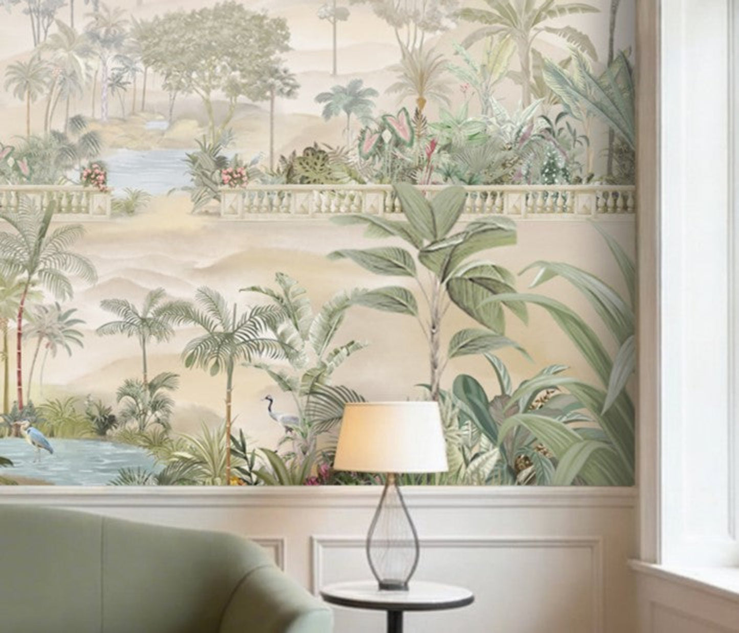 A spacious living room in soft beige tones featuring a large wall mural of palm trees, tropical plants, and a serene landscape with water and distant hills. The room includes a beige sofa with cushions, a green velvet sofa, two wooden armchairs, and a round marble coffee table. Warm pendant lights hang from the ceiling, and indoor plants complement the calm, neutral setting.