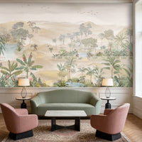 A spacious living room in soft beige tones featuring a large wall mural of palm trees, tropical plants, and a serene landscape with water and distant hills. The room includes a beige sofa with cushions, a green velvet sofa, two wooden armchairs, and a round marble coffee table. Warm pendant lights hang from the ceiling, and indoor plants complement the calm, neutral setting.