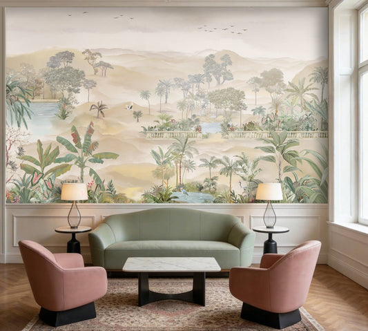 A spacious living room in soft beige tones featuring a large wall mural of palm trees, tropical plants, and a serene landscape with water and distant hills. The room includes a beige sofa with cushions, a green velvet sofa, two wooden armchairs, and a round marble coffee table. Warm pendant lights hang from the ceiling, and indoor plants complement the calm, neutral setting.