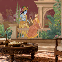 A large wall mural depicting the Hindu deity Krishna and the goddess Radha seated on a balcony, installed in a styled living room setting with a carved wooden coffee table and a wooden armchair in the foreground.