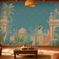 A large wall mural featuring an Indian classical scene with a blue peacock, a seated king and queen, palm trees, domes, and a teal background, shown installed in a warmly lit living room with a sofa, coffee table, and plants.