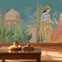 A large wall mural featuring an Indian classical scene with a blue peacock, a seated king and queen, palm trees, domes, and a teal background, shown installed in a warmly lit living room with a sofa, coffee table, and plants.