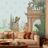 A styled living room featuring a large wall mural with a classical Indian scene, including a peacock, a woman in a blue sari, and architectural pillars, set behind a cream sectional sofa with decorative pillows.