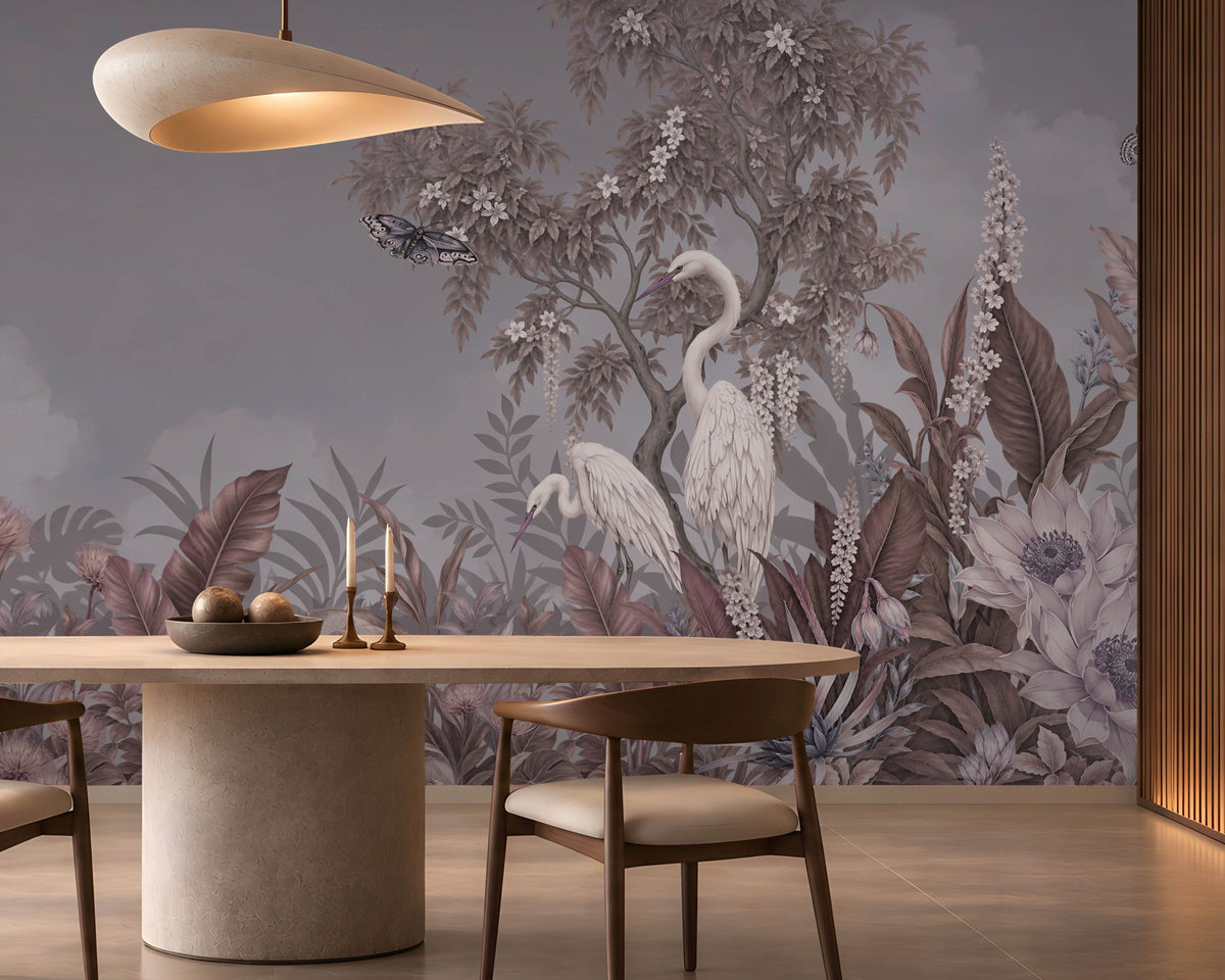 An elegant dining room featuring a large, scenic mural wallpaper illustration. The slate grey wallpaper depicts white herons standing amidst lush tropical foliage, flowering trees, and flying butterflies under a soft cloudy sky.