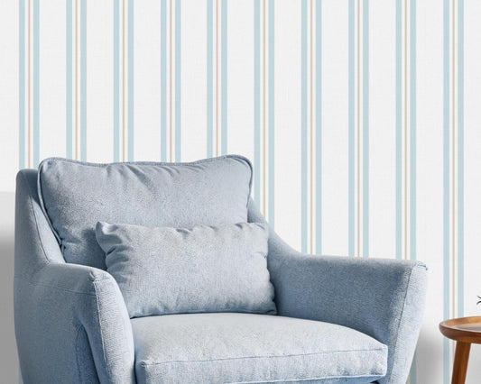 A bright sitting area features a clean coastal dune blue and white striped wallpaper.
The detailed design shows thin blue and beige vertical stripes repeating down a light base.
In front, a plush light blue armchair with a matching lumbar pillow rests comfortably.
The relaxing space is complete with a small wooden side table holding a mug and plant.