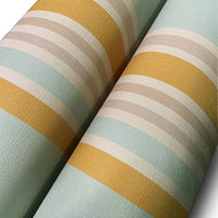 A child's playroom features a muted olive green wallpaper with warm colored stripes.
The detailed pattern shows vertical bands of orange, yellow, and cream running down the wall.
In front, a white giraffe-themed dresser stands next to a dark rocking chair and pouf.
The playful space is neatly arranged with children's toys and a round world map rug.