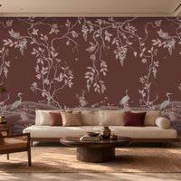Timeless Avian Garden Mural in rich burgundy brown featuring delicate pink botanical branches, graceful birds, soft floral details, and flowing foliage. The elegant nature-inspired wallpaper creates a warm and sophisticated living room with neutral seating, dark wood furniture, natural textures, and soft ambient lighting.
