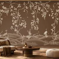 Warm brown botanical wallpaper mural featuring delicate ivory vines, leaves, blossoms, and elegant birds, styled in a luxurious living room with natural wood furniture, a cane accent chair, soft neutral seating, a textured rug, warm ambient lighting, and refined earthy décor.
