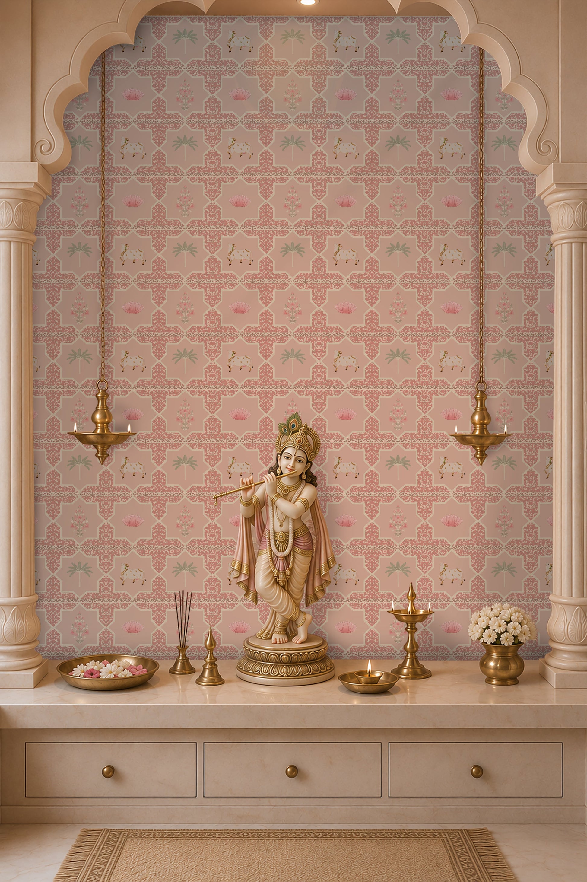 A beautifully styled Indian mandir backdrop or pooja room altar framed by architectural pillars, featuring a pink Vraj Kamal Pichwai wallpaper with repeating cow and lotus motifs behind a standing Lord Krishna idol and brass lamps.