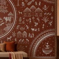 Warli Divine Harmony - Burnt Terracotta Red wallpaper featuring intricate white Warli tribal artwork with a central Radha Krishna motif, traditional village scenes, dancing figures, trees, huts, and decorative borders. Set in a warm Indian heritage-inspired living room with carved wooden furniture, earthy textiles, and elegant terracotta tones. 
