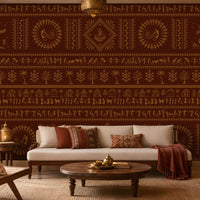 A traditional Indian living room titled “Warli Folk Tales – Vintage Reddish Brown.” The focal wall is covered in deep reddish-brown wallpaper with intricate golden tribal art depicting dancing figures, farming scenes, huts, animals, and trees arranged in horizontal bands. A beige upholstered sofa with patterned earthy cushions sits beside a woven chair with a floral cushion, centered around a round wooden coffee table adorned with brass decorative items. Warm lighting from a brass hanging lamp and table.