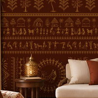 A traditional Indian living room titled “Warli Folk Tales – Vintage Reddish Brown.” The focal wall is covered in deep reddish-brown wallpaper with intricate golden tribal art depicting dancing figures, farming scenes, huts, animals, and trees arranged in horizontal bands. A beige upholstered sofa with patterned earthy cushions sits beside a woven chair with a floral cushion, centered around a round wooden coffee table adorned with brass decorative items. Warm lighting from a brass hanging lamp and table.