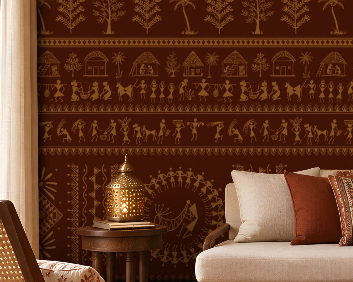 A traditional Indian living room titled “Warli Folk Tales – Vintage Reddish Brown.” The focal wall is covered in deep reddish-brown wallpaper with intricate golden tribal art depicting dancing figures, farming scenes, huts, animals, and trees arranged in horizontal bands. A beige upholstered sofa with patterned earthy cushions sits beside a woven chair with a floral cushion, centered around a round wooden coffee table adorned with brass decorative items. Warm lighting from a brass hanging lamp and table.