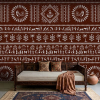 A traditional Indian living room titled “Warli Folk Tales – Vintage Reddish Brown.” The focal wall is painted in a deep reddish-brown shade and decorated with intricate white Warli tribal art depicting village life, including dancing figures, farming scenes, animals, trees, and geometric motifs. A carved wooden daybed with patterned cushions and a brown throw blanket sits against the wall, accompanied by a clay pot on the floor and a woven rug with geometric designs. The warm, earthy palette and handcrafted