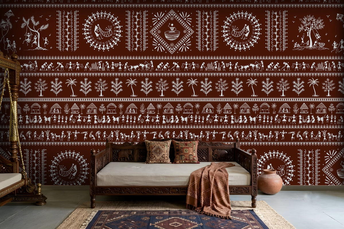 A traditional Indian living room titled “Warli Folk Tales – Vintage Reddish Brown.” The focal wall is painted in a deep reddish-brown shade and decorated with intricate white Warli tribal art depicting village life, including dancing figures, farming scenes, animals, trees, and geometric motifs. A carved wooden daybed with patterned cushions and a brown throw blanket sits against the wall, accompanied by a clay pot on the floor and a woven rug with geometric designs. The warm, earthy palette and handcrafted
