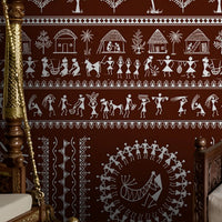 A traditional Indian living room titled “Warli Folk Tales – Vintage Reddish Brown.” The focal wall is painted in a deep reddish-brown shade and decorated with intricate white Warli tribal art depicting village life, including dancing figures, farming scenes, animals, trees, and geometric motifs. A carved wooden daybed with patterned cushions and a brown throw blanket sits against the wall, accompanied by a clay pot on the floor and a woven rug with geometric designs. The warm, earthy palette and handcrafted