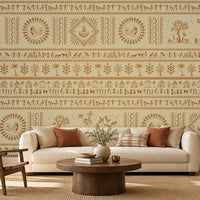 A contemporary Indian living room titled “Warli Folk Tales – Vintage Beige Brown.” The focal wall is painted beige and decorated with intricate brown Warli tribal art featuring human figures, animals, huts, trees, and geometric motifs arranged in horizontal bands. A white sofa with beige and rust-colored cushions sits beside a wooden armchair with a woven seat, centered around a round wooden coffee table with decorative items. A side table with a lamp and potted plants add warmth and greenery, while natural