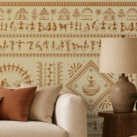 A contemporary Indian living room titled “Warli Folk Tales – Vintage Beige Brown.” The focal wall is painted beige and decorated with intricate brown Warli tribal art featuring human figures, animals, huts, trees, and geometric motifs arranged in horizontal bands. A white sofa with beige and rust-colored cushions sits beside a wooden armchair with a woven seat, centered around a round wooden coffee table with decorative items. A side table with a lamp and potted plants add warmth and greenery, while natural
