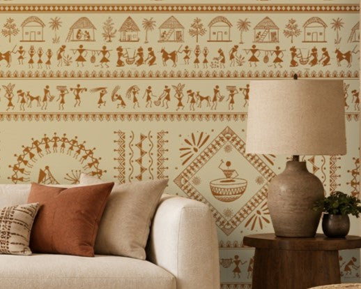 A contemporary Indian living room titled “Warli Folk Tales – Vintage Beige Brown.” The focal wall is painted beige and decorated with intricate brown Warli tribal art featuring human figures, animals, huts, trees, and geometric motifs arranged in horizontal bands. A white sofa with beige and rust-colored cushions sits beside a wooden armchair with a woven seat, centered around a round wooden coffee table with decorative items. A side table with a lamp and potted plants add warmth and greenery, while natural