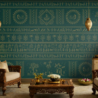 A traditional Indian living room titled “Warli Folk Tales – Vintage Teal Green.” The focal wall is painted in a deep teal shade and adorned with golden Warli tribal art depicting village life, dancing figures, huts, and trees in horizontal bands. Two intricately carved wooden armchairs with cushions flank a central coffee table decorated with brass items and a bowl of white flowers. Hanging brass pendant lamps and a golden table lamp cast a warm glow. 