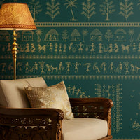A traditional Indian living room titled “Warli Folk Tales – Vintage Teal Green.” The focal wall is painted in a deep teal shade and adorned with golden Warli tribal art depicting village life, dancing figures, huts, and trees in horizontal bands. Two intricately carved wooden armchairs with cushions flank a central coffee table decorated with brass items and a bowl of white flowers. Hanging brass pendant lamps and a golden table lamp cast a warm glow.