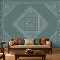 A styled living room featuring a large teal wall mural with intricate white Indian folk art patterns, including a central diamond mandala. The room is furnished with a cream-colored sofa decorated with patterned cushions, a dark wood coffee table, and traditional wooden furniture.
