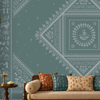 A styled living room featuring a large teal wall mural with intricate white Indian folk art patterns, including a central diamond mandala. The room is furnished with a cream-colored sofa decorated with patterned cushions, a dark wood coffee table, and traditional wooden furniture.