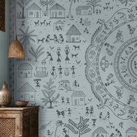 A large wall mural featuring a detailed blue-gray circular mandala and surrounding folk-art style illustrations of houses, people, and plants, shown in a styled living room with wooden furniture and woven decor.