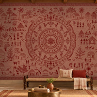 A large wall mural featuring a detailed red and dusty rose Indian folk art style mandala with village scenes, surrounded by a furnished room with a bench, pillows, plants, and a brass table.