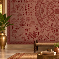 A large wall mural featuring a detailed red and dusty rose Indian folk art style mandala with village scenes, surrounded by a furnished room with a bench, pillows, plants, and a brass table.