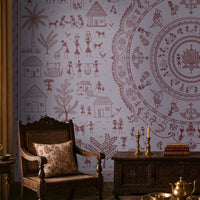 A styled traditional living room featuring a large lavender wall mural with a detailed central mandala and surrounding folk-art illustrations in muted red-brown. The room is furnished with dark wood furniture, patterned cushions, a brass table lamp, and a patterned area rug.