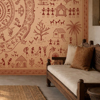 A styled interior scene featuring a large terracotta-colored wall mural with traditional Indian folk art illustrations, including figures, animals, houses, and motifs, paired with a carved wooden sofa and cushions in a warm, rustic room.