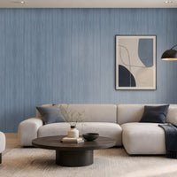 Elegant modern living room with soft blue wood-textured wallpaper covering the main feature wall, complemented by a large cream sectional sofa, matching armchair, dark round coffee table, neutral textured rug, and contemporary abstract artwork. Floor-to-ceiling windows with sheer curtains allow natural daylight to illuminate the calm, sophisticated interior, creating a serene and refined atmosphere.
