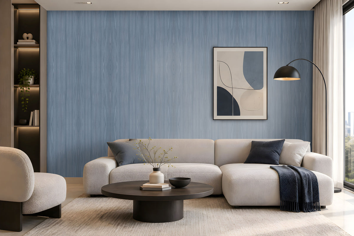 Elegant modern living room with soft blue wood-textured wallpaper covering the main feature wall, complemented by a large cream sectional sofa, matching armchair, dark round coffee table, neutral textured rug, and contemporary abstract artwork. Floor-to-ceiling windows with sheer curtains allow natural daylight to illuminate the calm, sophisticated interior, creating a serene and refined atmosphere.
