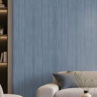 Elegant modern living room with soft blue wood-textured wallpaper covering the main feature wall, complemented by a large cream sectional sofa, matching armchair, dark round coffee table, neutral textured rug, and contemporary abstract artwork. Floor-to-ceiling windows with sheer curtains allow natural daylight to illuminate the calm, sophisticated interior, creating a serene and refined atmosphere.