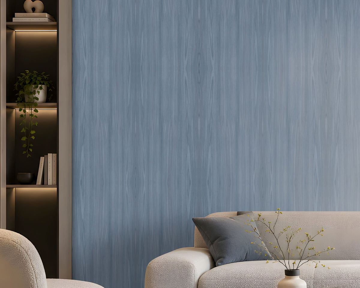 Elegant modern living room with soft blue wood-textured wallpaper covering the main feature wall, complemented by a large cream sectional sofa, matching armchair, dark round coffee table, neutral textured rug, and contemporary abstract artwork. Floor-to-ceiling windows with sheer curtains allow natural daylight to illuminate the calm, sophisticated interior, creating a serene and refined atmosphere.