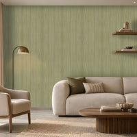 Modern living room with a soft green textured accent wall, cream upholstered sofa and armchair, warm floor lamp, floating wooden shelves with books and plants, and a round wooden coffee table on a neutral area rug. Natural light enters through a large window with beige curtains, creating a calm, contemporary interior.