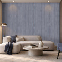 Elegant contemporary living room featuring a soft blue-grey wood-textured wallpaper across the main feature wall, paired with a spacious cream boucle sectional sofa, blue and neutral accent cushions, and a dark blue throw. A round light stone coffee table with stacked books and a decorative vase sits on a textured neutral rug, while a sculptural floor lamp, built-in illuminated shelving, and large windows bring warmth and natural light to the sophisticated space.