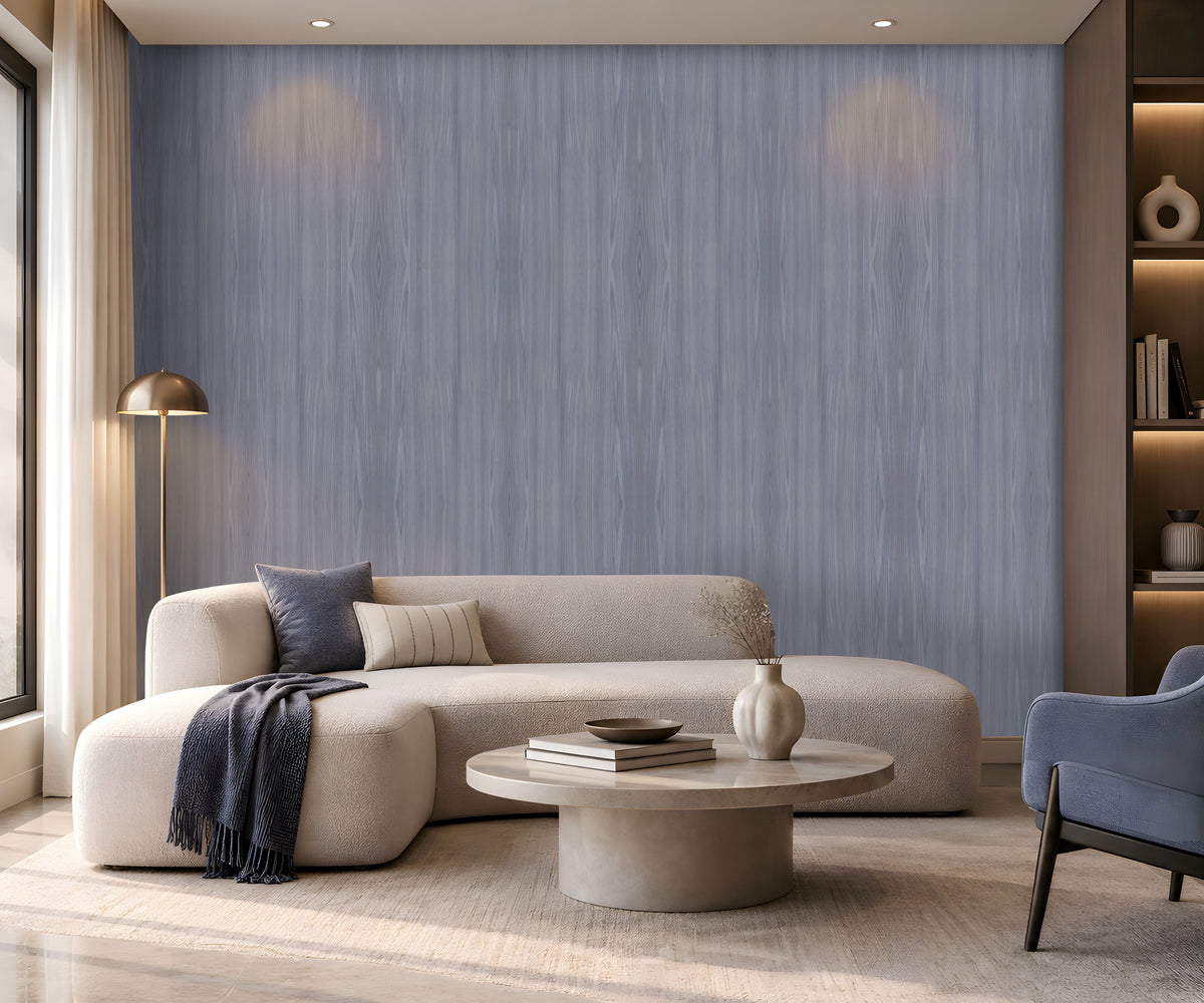 Elegant contemporary living room featuring a soft blue-grey wood-textured wallpaper across the main feature wall, paired with a spacious cream boucle sectional sofa, blue and neutral accent cushions, and a dark blue throw. A round light stone coffee table with stacked books and a decorative vase sits on a textured neutral rug, while a sculptural floor lamp, built-in illuminated shelving, and large windows bring warmth and natural light to the sophisticated space.