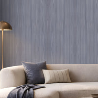 Elegant contemporary living room featuring a soft blue-grey wood-textured wallpaper across the main feature wall, paired with a spacious cream boucle sectional sofa, blue and neutral accent cushions, and a dark blue throw. A round light stone coffee table with stacked books and a decorative vase sits on a textured neutral rug, while a sculptural floor lamp, built-in illuminated shelving, and large windows bring warmth and natural light to the sophisticated space.
