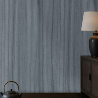 Wood Seamless Wallpaper - Stone Blue featured in a Japandi-inspired living space. The wallpaper showcases a seamless vertical wood grain texture in soft ash grey tones, creating a sophisticated and calming backdrop. Paired with dark wood furniture, built-in illuminated shelving, neutral floor cushions, and warm ambient lighting, the design adds depth, elegance, and a contemporary natural aesthetic to the interior.