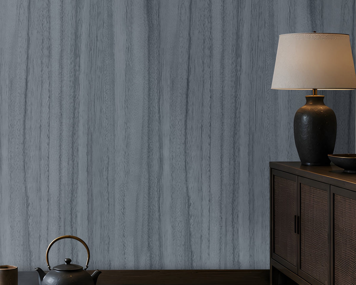 Wood Seamless Wallpaper - Stone Blue featured in a Japandi-inspired living space. The wallpaper showcases a seamless vertical wood grain texture in soft ash grey tones, creating a sophisticated and calming backdrop. Paired with dark wood furniture, built-in illuminated shelving, neutral floor cushions, and warm ambient lighting, the design adds depth, elegance, and a contemporary natural aesthetic to the interior.