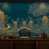 Luxurious home office featuring a heritage-inspired mural in rich peacock blue and antique gold. The mural depicts ornate domed pavilions, lush botanical foliage, an elegant arched bridge, and graceful swans on a tranquil pond, with subtle palace architecture in the background. The space is styled with a carved dark wood executive desk, a tufted leather chair, brass desk accessories, a vintage clock, an adjustable green banker’s lamp, built-in bookshelves with decorative objects, and warm ambient lighting, 